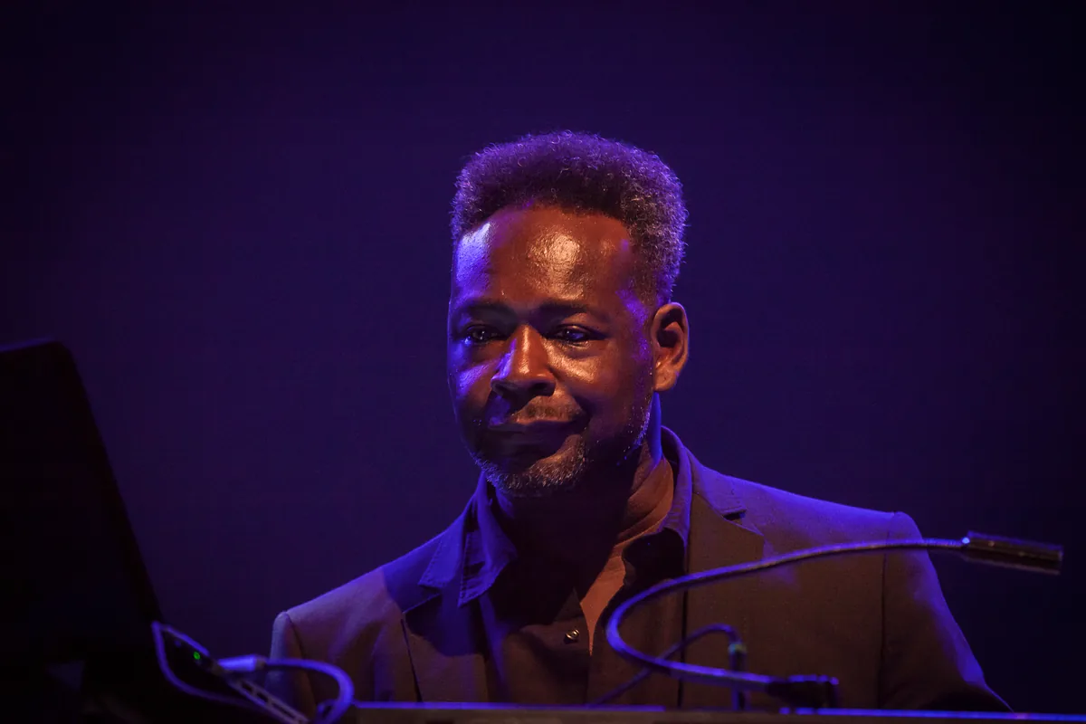 A Guy Called Gerald behind his equipment on stage under purple light