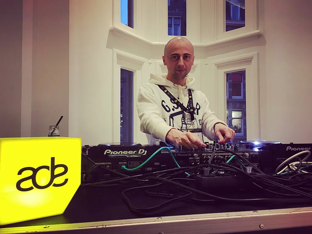 A DJ at a Pioneer DJ setup beside a yellow illuminated ADE sign at Amsterdam Dance Event