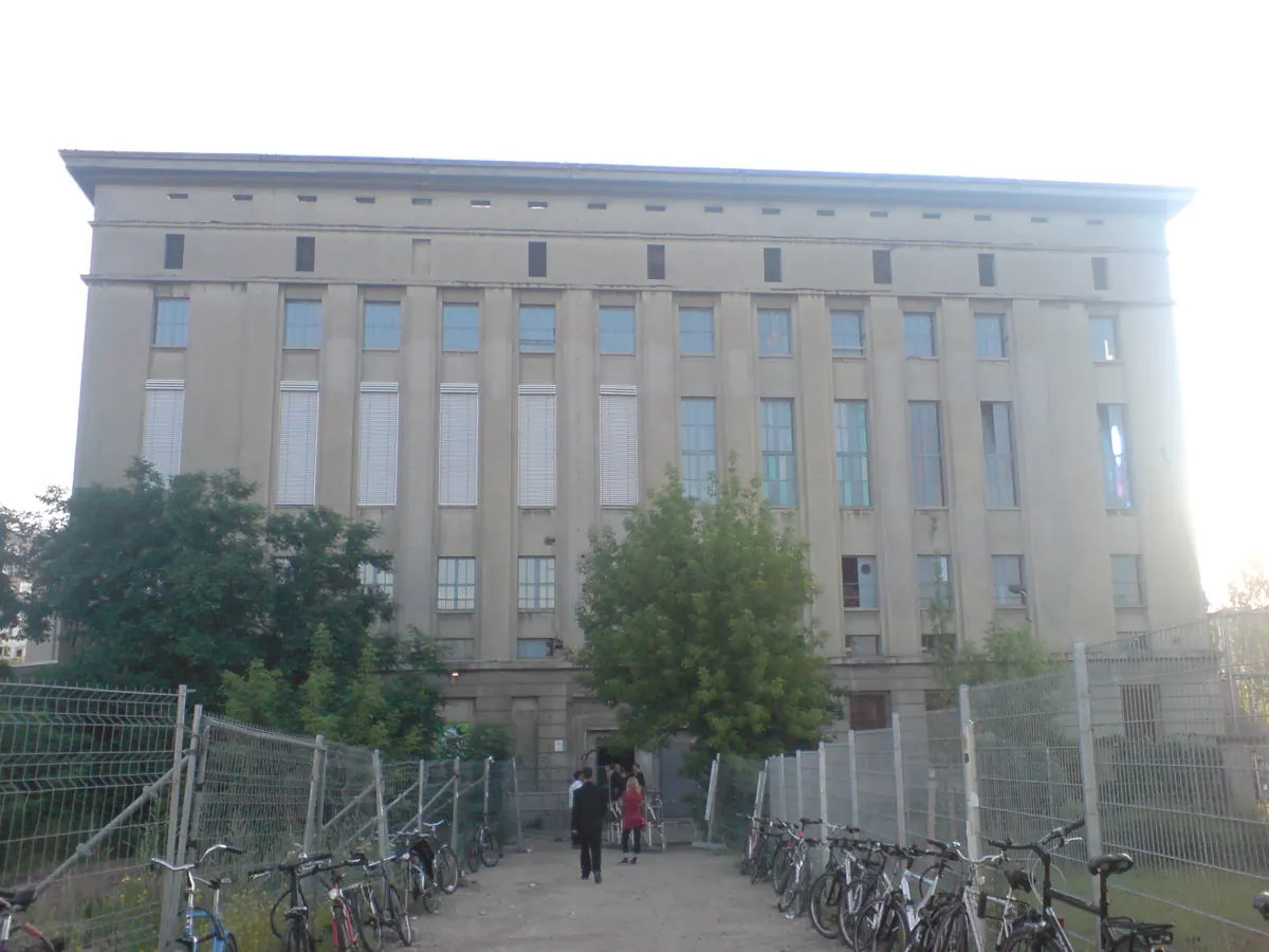 The grey neoclassical front of the Berghain building in Berlin, with a few people at the entrance and bicycles parked behind fences