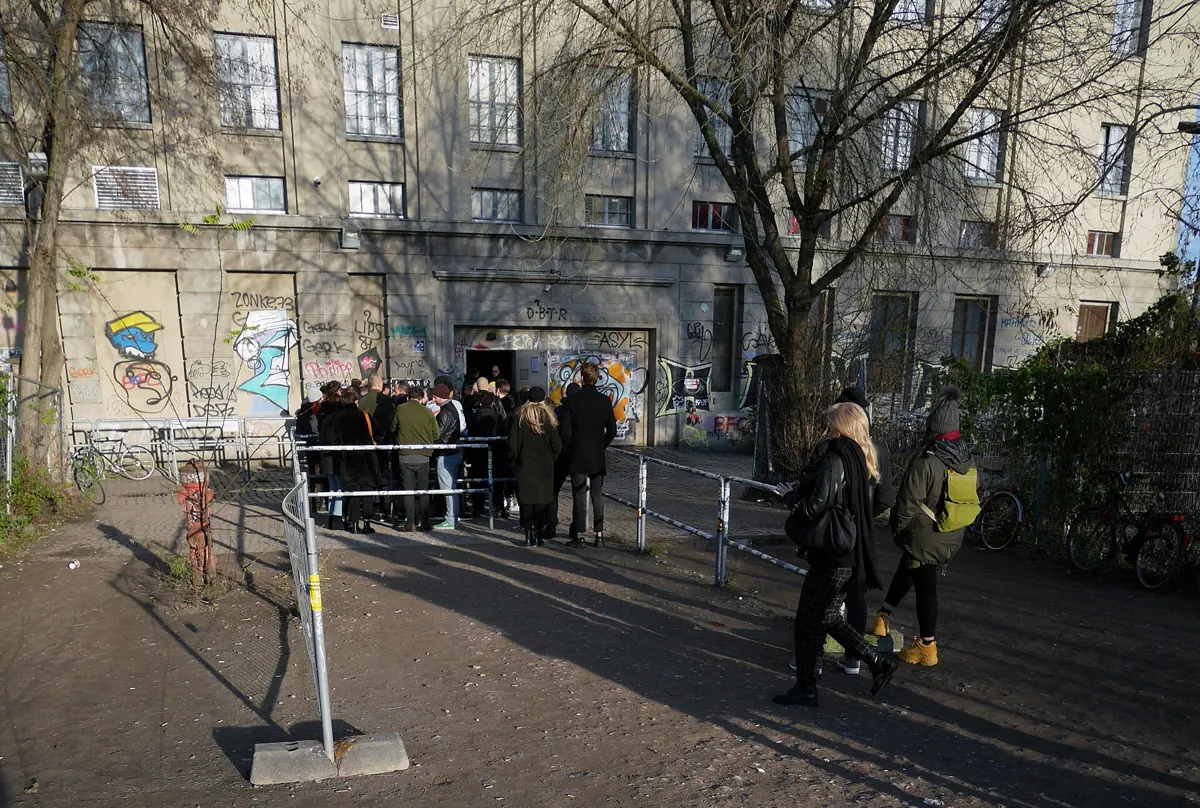 People standing behind metal barriers in front of the Berghain entrance, which is covered in graffiti