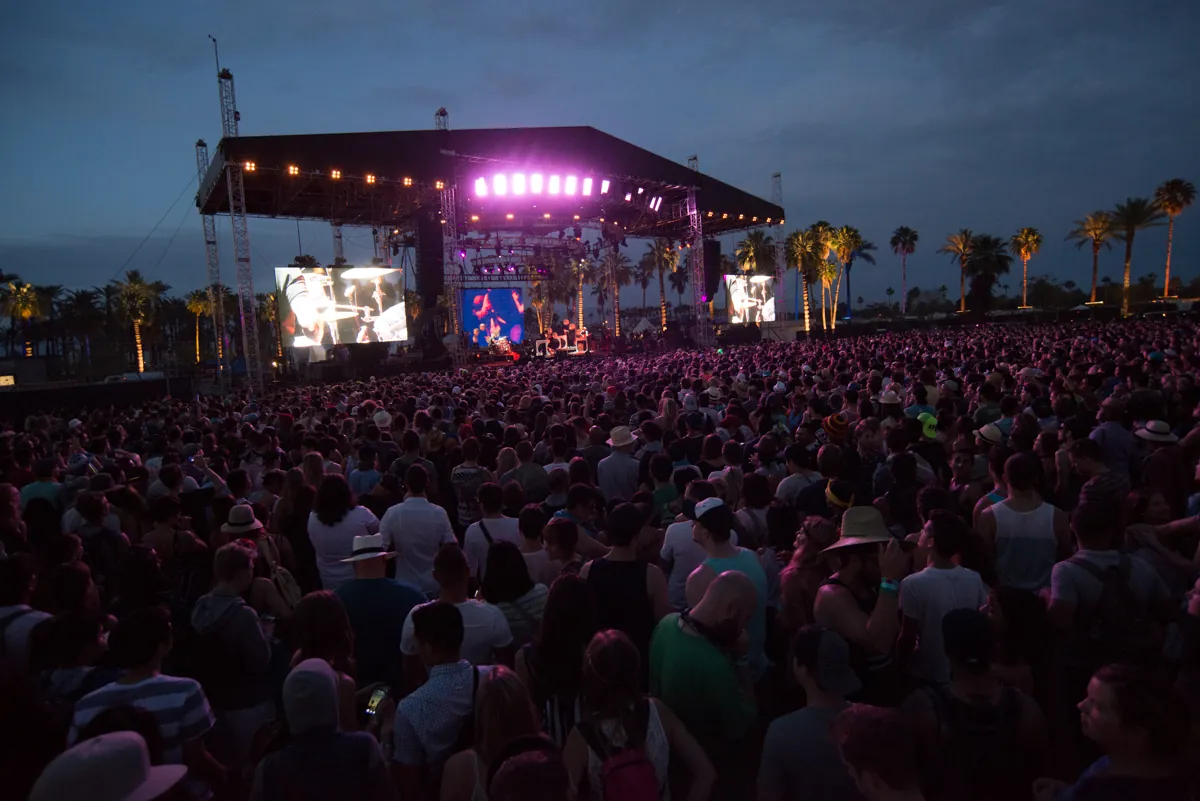 A large crowd facing the Outdoor Theatre stage at dusk in 2014, screens either side of the stage and palm trees along the edge of the field