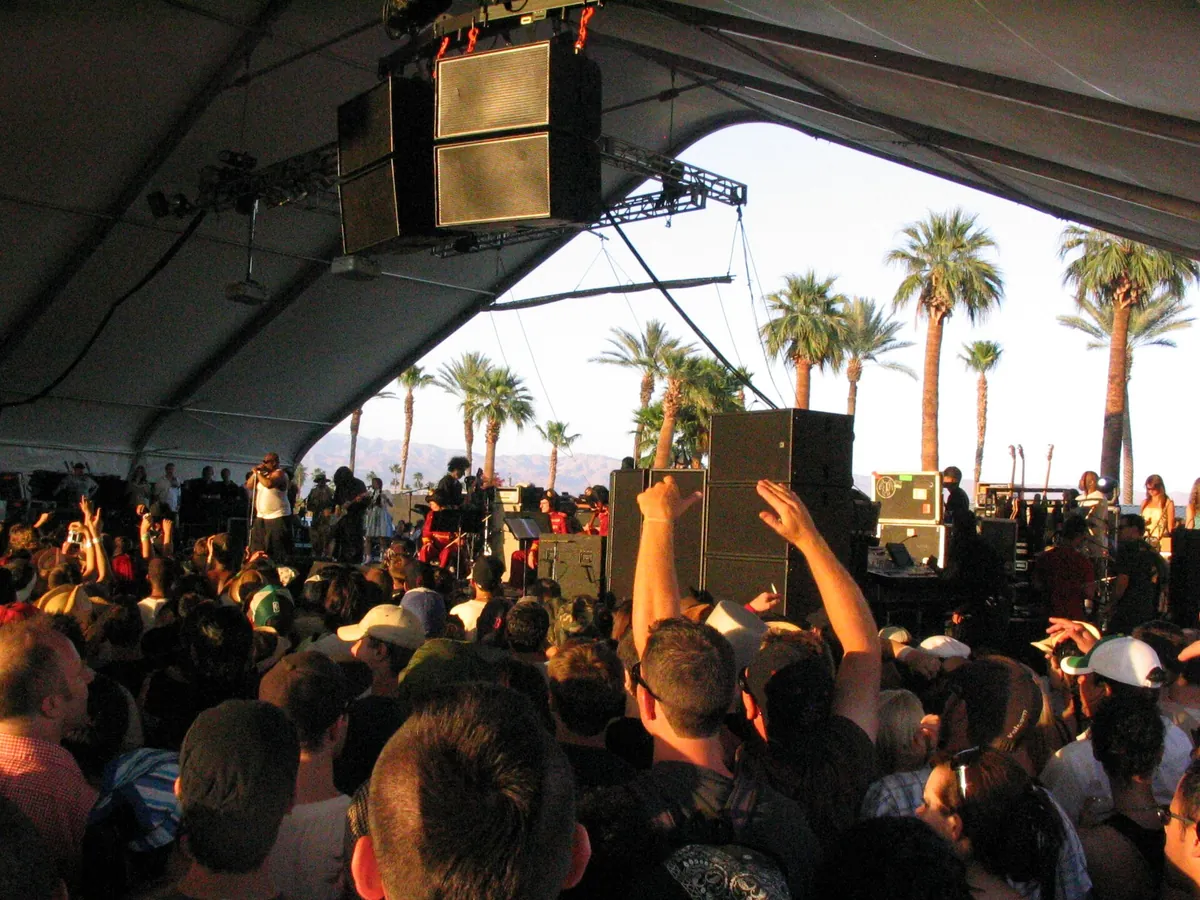 A crowd under a white festival tent at Coachella in 2006, a band on the stage, speakers hanging from the roof and palm trees in evening light beyond the open side