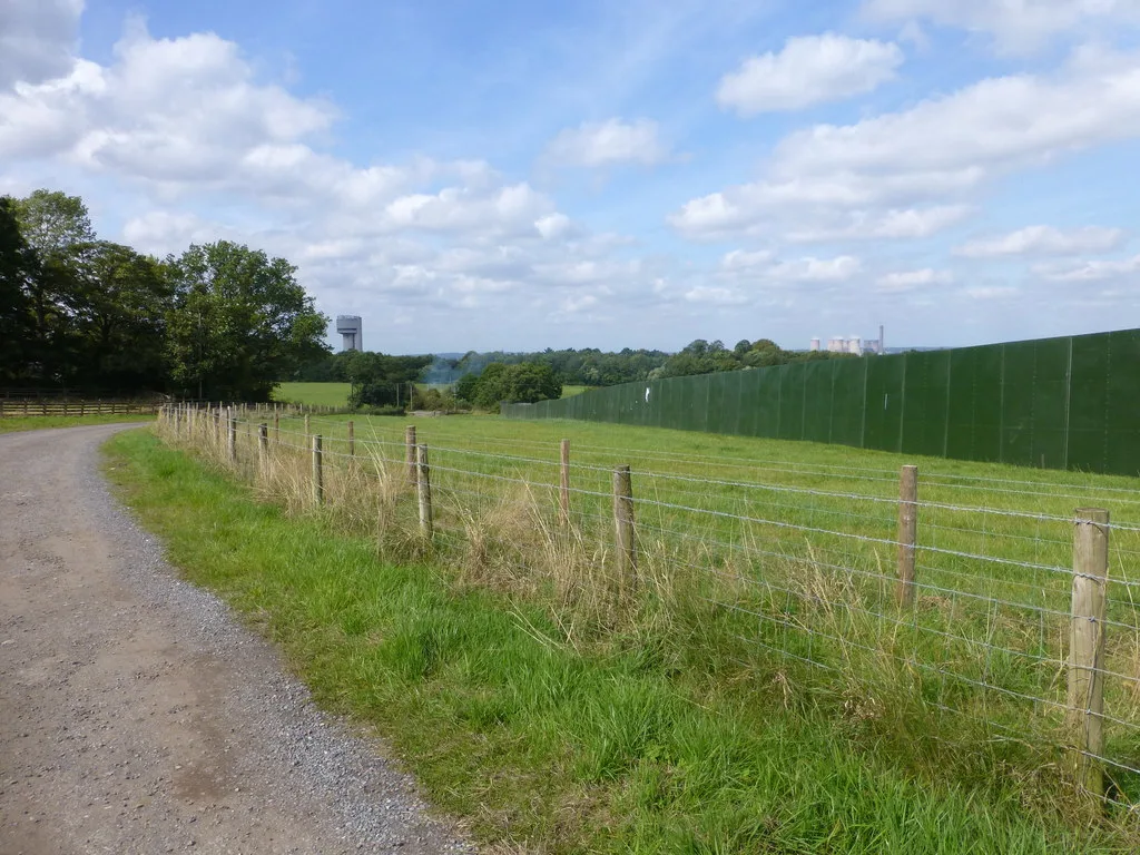 A farm track beside green fencing at the edge of the Creamfields site near Daresbury, with fields and trees beyond