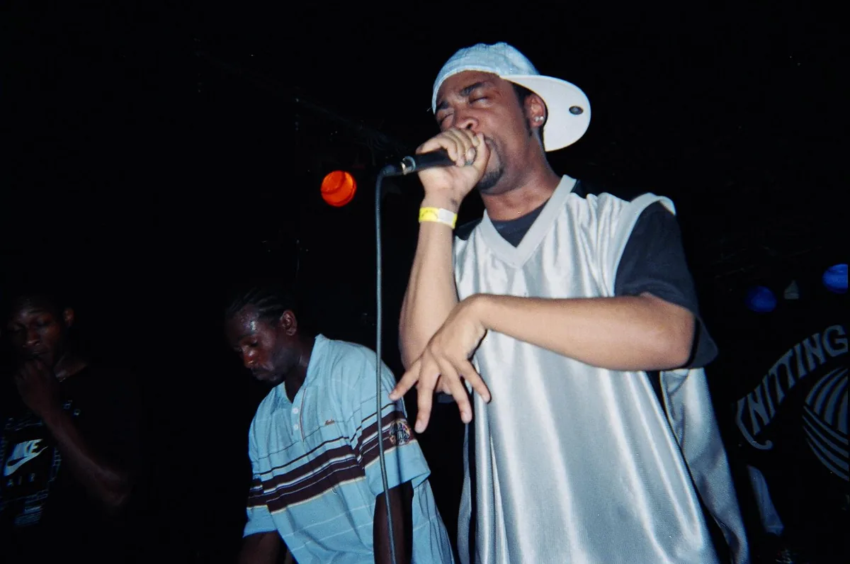 Two Roll Deep MCs on a dark stage in 2005, one holding a microphone close in a white cap and silver vest