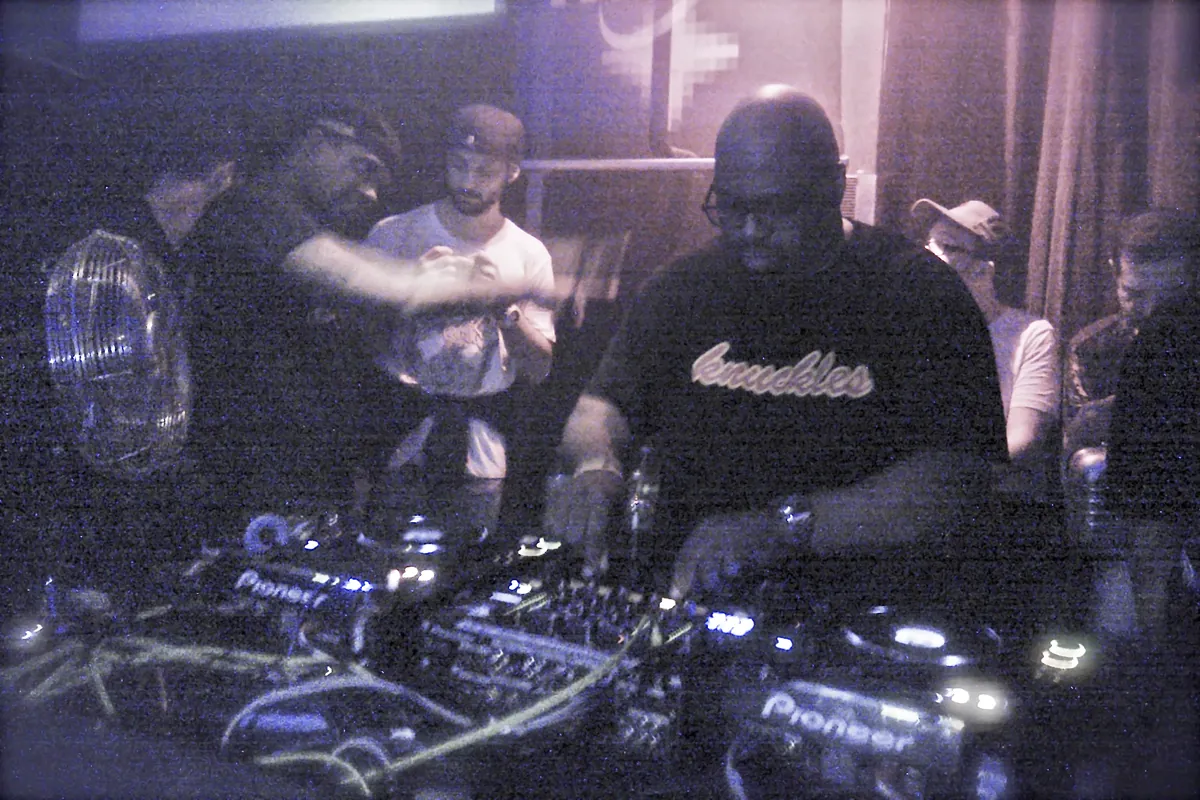 Frankie Knuckles behind the decks at a club in Amsterdam, lit in purple, with people crowding the booth
