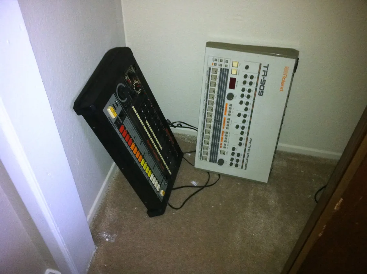 A Roland TR-808 drum machine leaning against a wall beside a Roland TR-909 on a studio floor