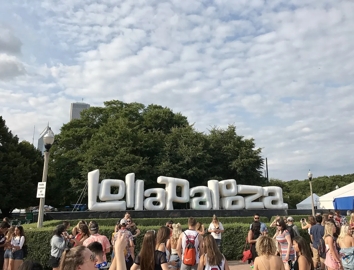 The word Lollapalooza in giant white inflatable letters in front of trees and a skyscraper, festivalgoers walking past in the foreground