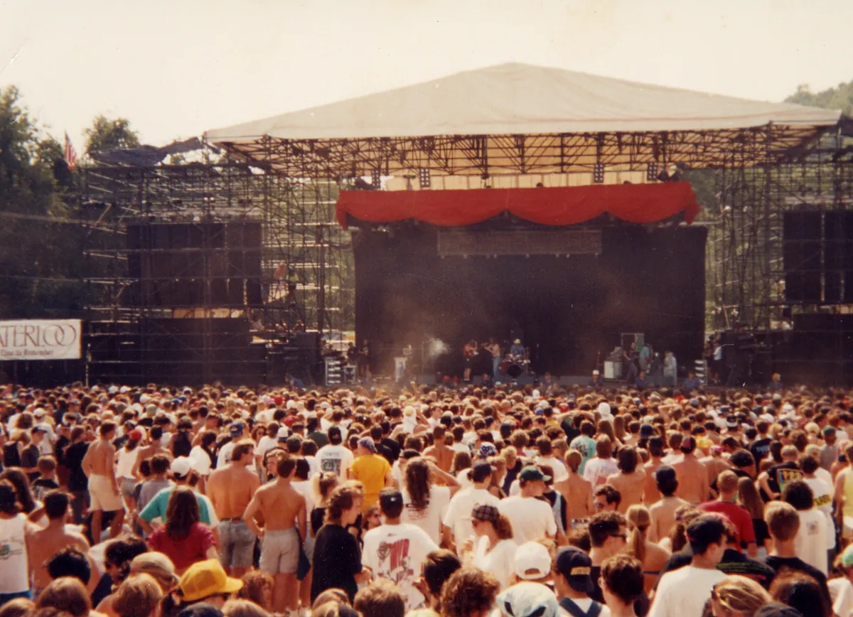 A large outdoor crowd facing a scaffold stage with a red-draped roof on the first Lollapalooza tour in 1991