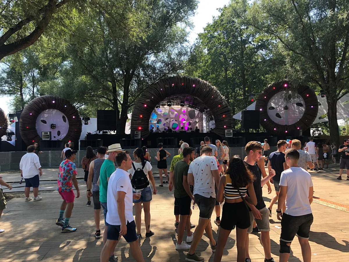 A stage under tall trees framed by three giant woven rings, with people dancing on a wooden floor in the sun