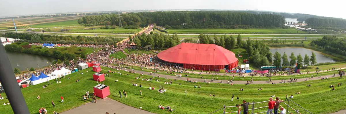 A wide panorama over green festival grounds with a large red big-top tent, crowds on paths, ponds and a line of trees beyond