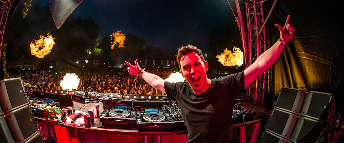 Hardwell at the decks with both arms raised, flames bursting behind him and a night crowd beyond