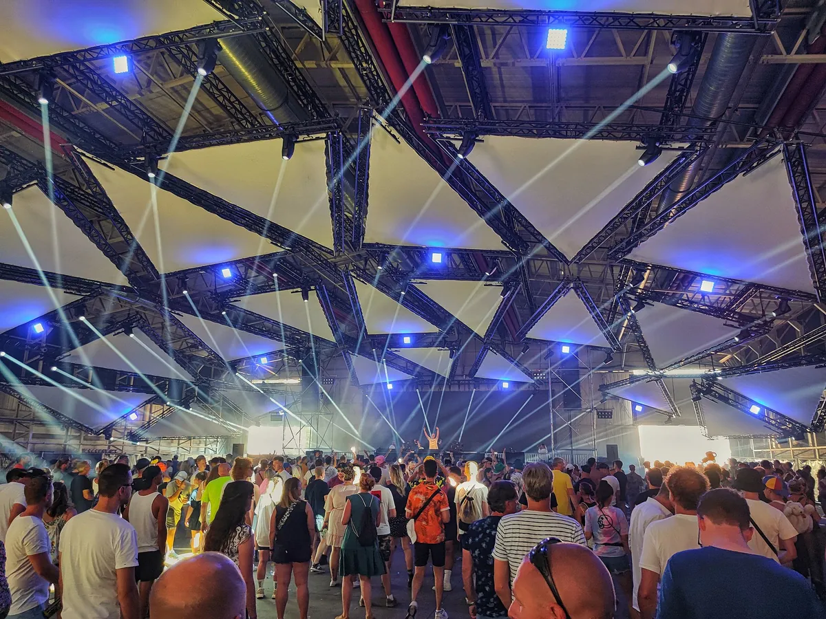 A packed crowd under a lattice roof lit by blue and white beams inside the Cloud Factory hangar in 2022