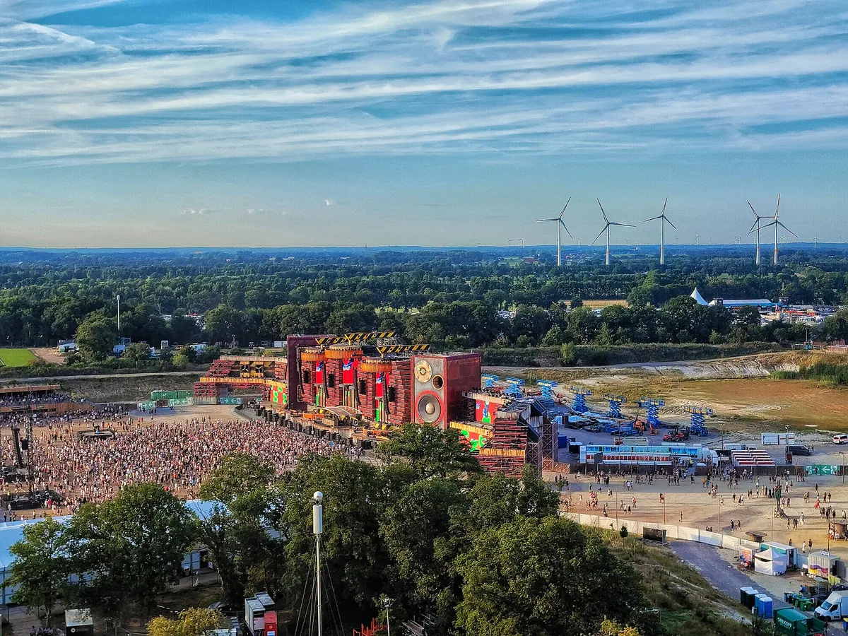 The Parookaville Mainstage from the air in 2022, a red and gold stage with the crowd in front of it, tents and fields beyond and wind turbines on the horizon