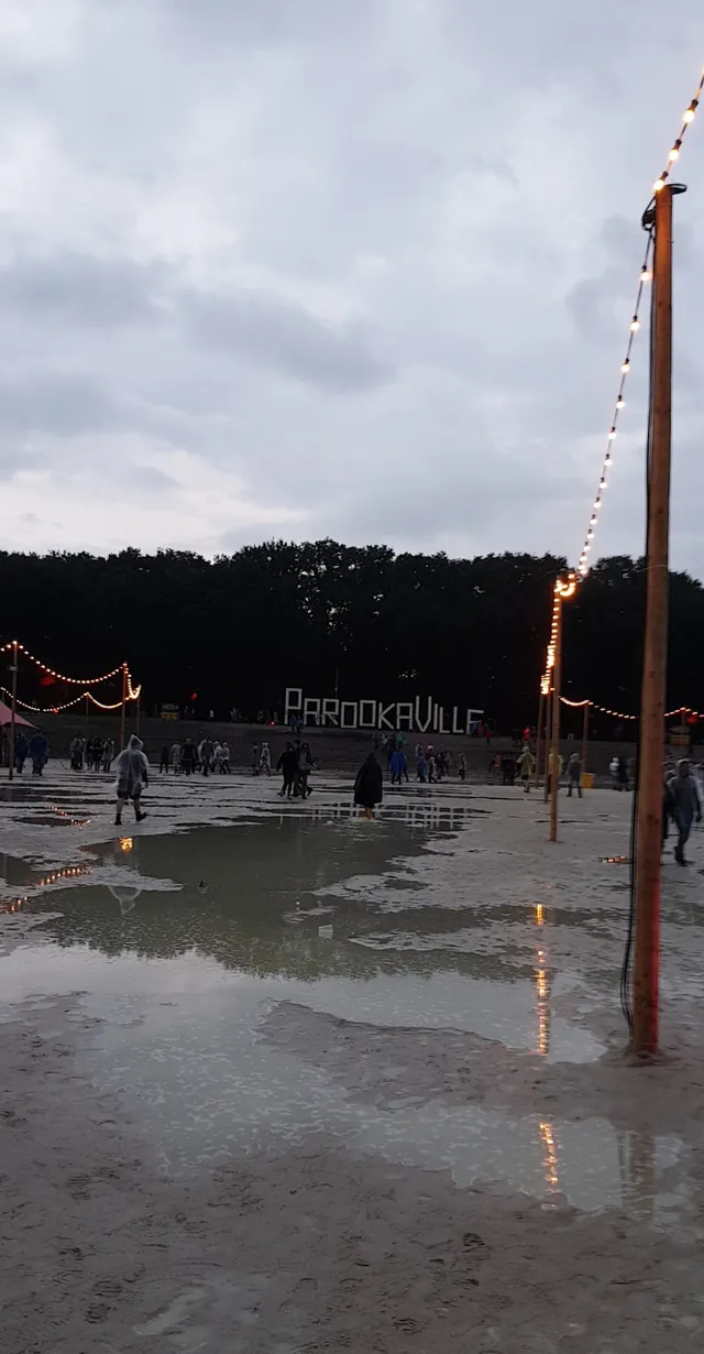 A flooded festival ground at dusk, with the PAROOKAVILLE letters beyond the water and strings of lights on poles