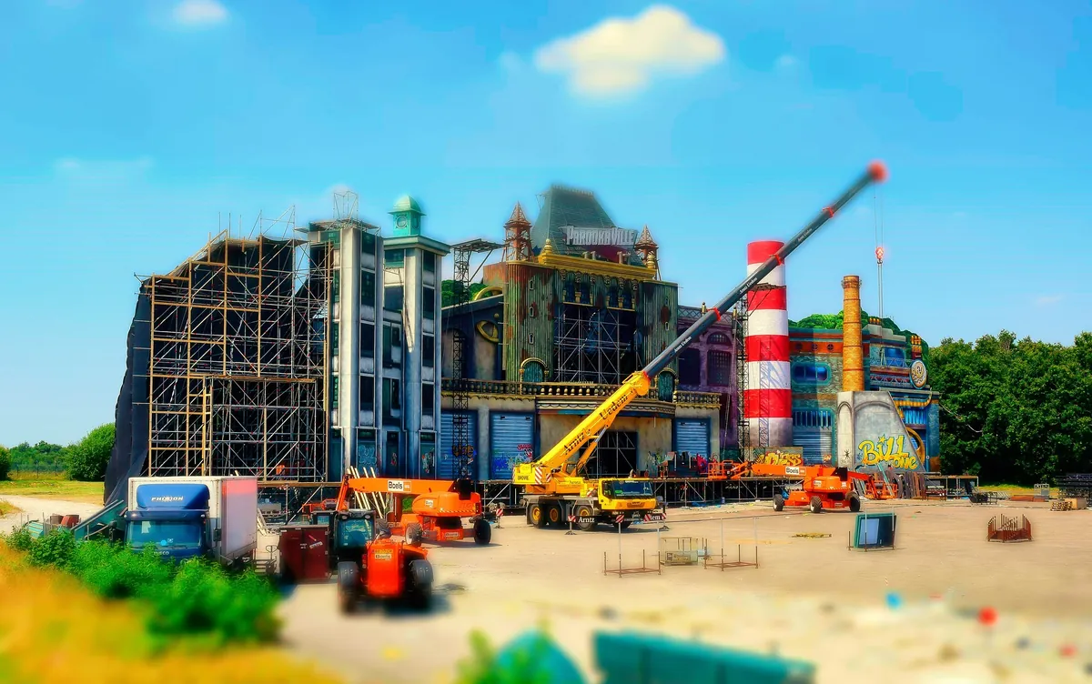 A stage facade of mock town buildings with scaffolding and a red and white chimney, cranes and forklifts in front, on the Parookaville site in 2016