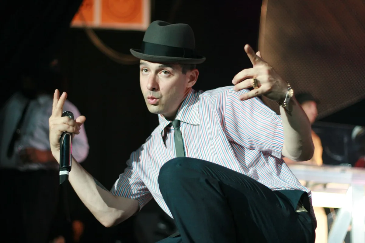 Ad-Rock of the Beastie Boys in a grey fedora and striped shirt, crouching with a microphone on stage