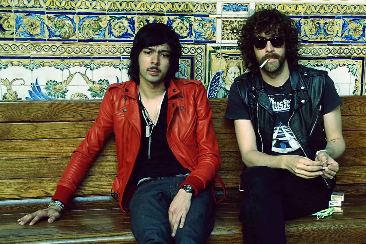 The two members of Justice sitting on a wooden bench in front of a wall of painted Spanish tiles