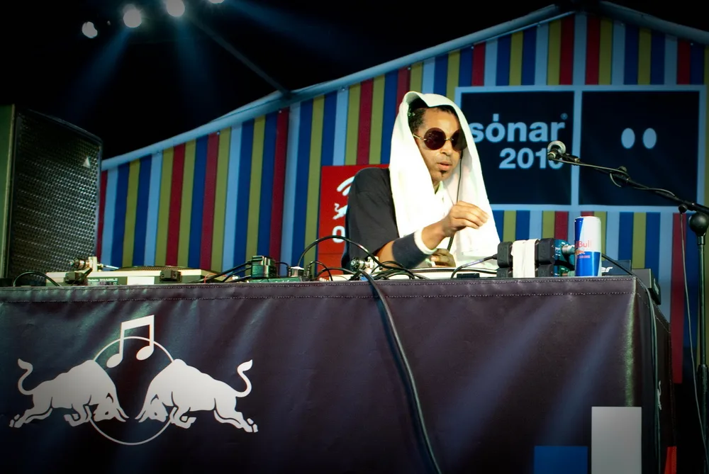 Moodymann in sunglasses and a white head covering at a DJ table, with a Sónar 2010 banner behind him