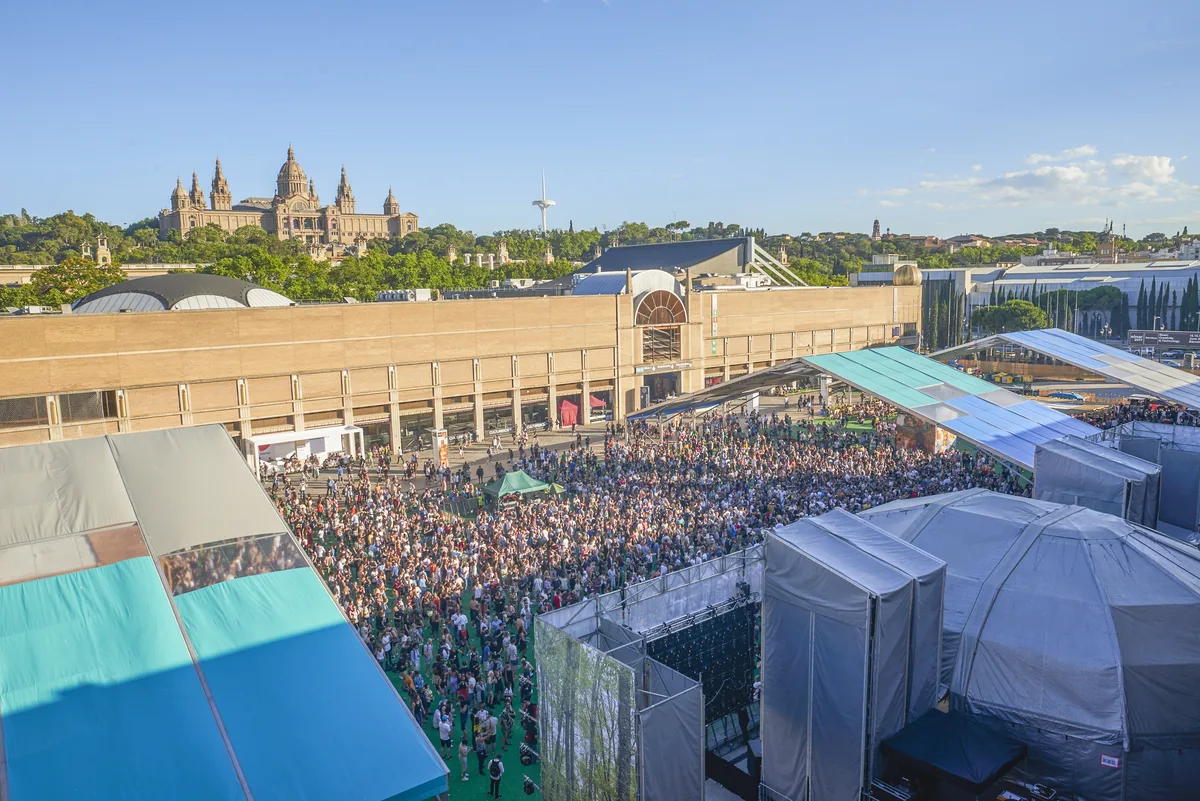 A crowd filling the open-air SonarVillage stage at Fira Montjuïc in afternoon sun, with the domed Palau Nacional on the hill behind