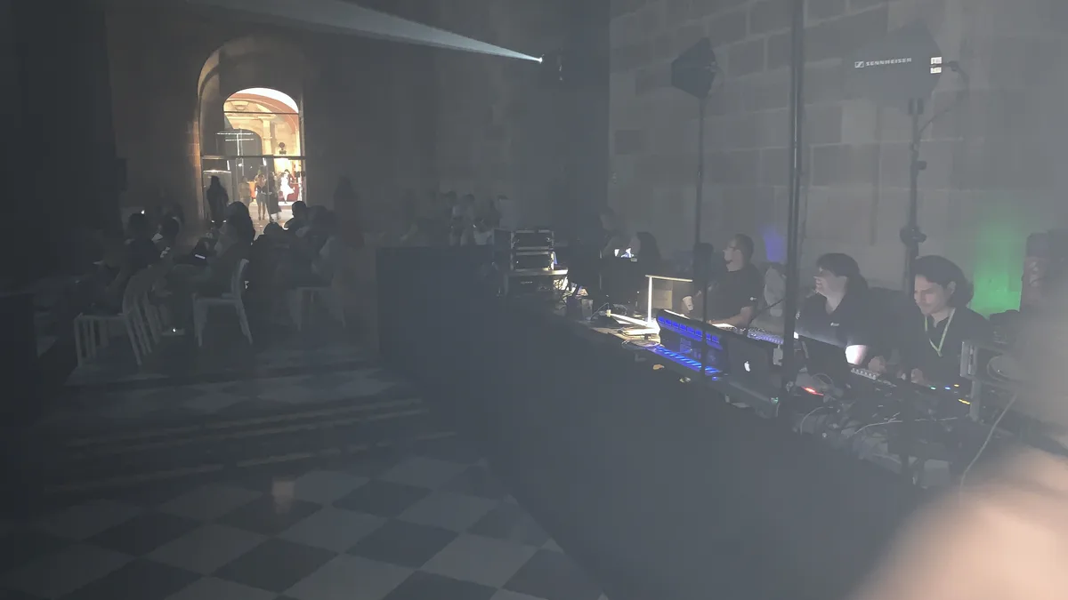 A dim stone hall with an arched doorway and a chequered floor, with people at laptops and mixing desks along one side
