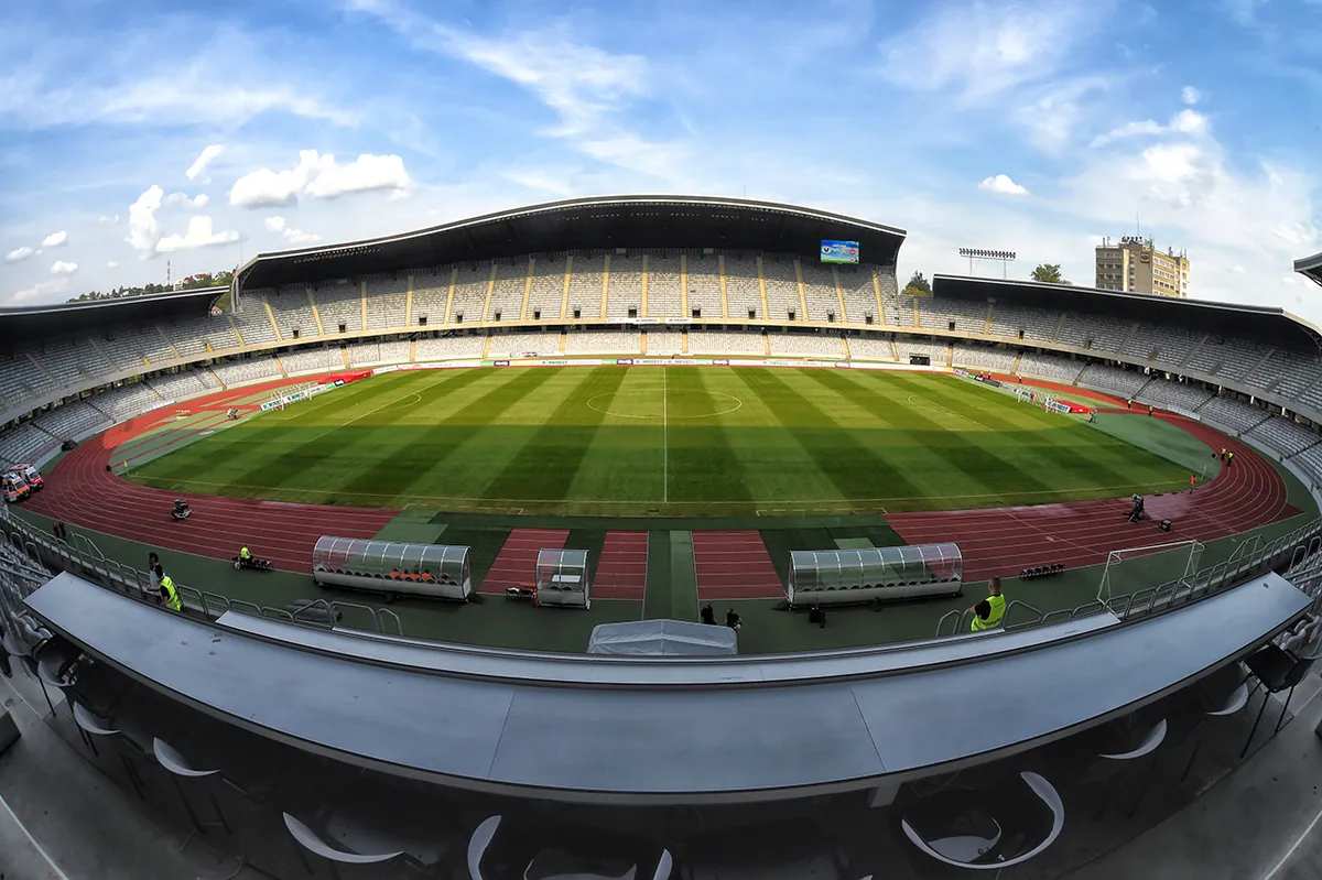 Cluj Arena from inside, an oval football stadium with a curved roof over grey seats, a green pitch and a running track, under a blue sky