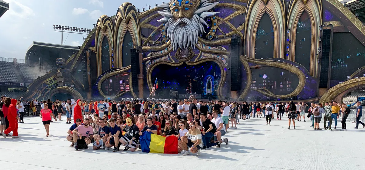 A group of festivalgoers sitting and standing with a Romanian flag in front of the bearded-wizard main stage at Untold 2019, in daylight
