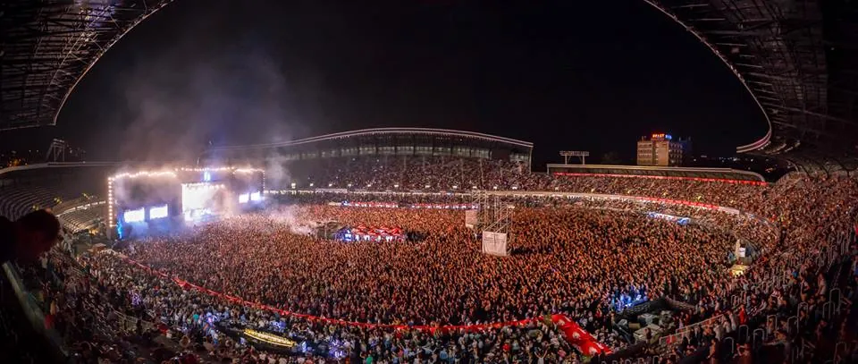 A wide night panorama of Cluj Arena at Untold 2015, the pitch and every stand packed, the lit main stage at the far left