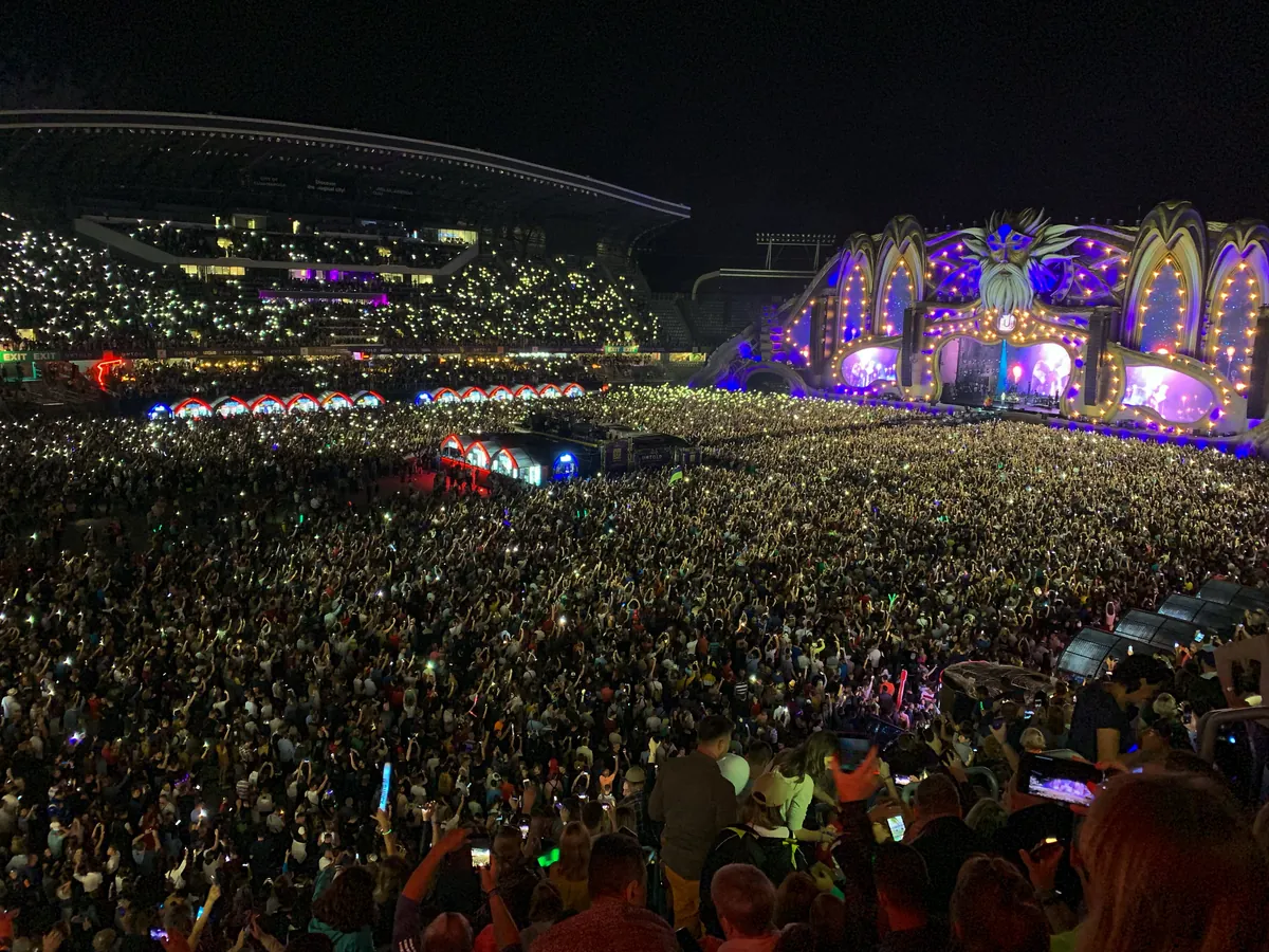 A packed crowd holding up phone lights in front of the purple and gold Untold main stage at night in 2019, the stadium stands full behind
