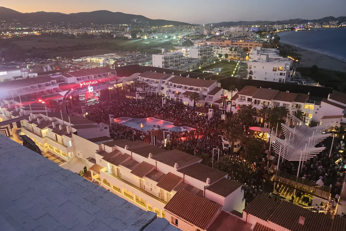 The Ushuaia Ibiza dance floor packed around the pool at dusk, seen from a high window, with the hills and the bay behind