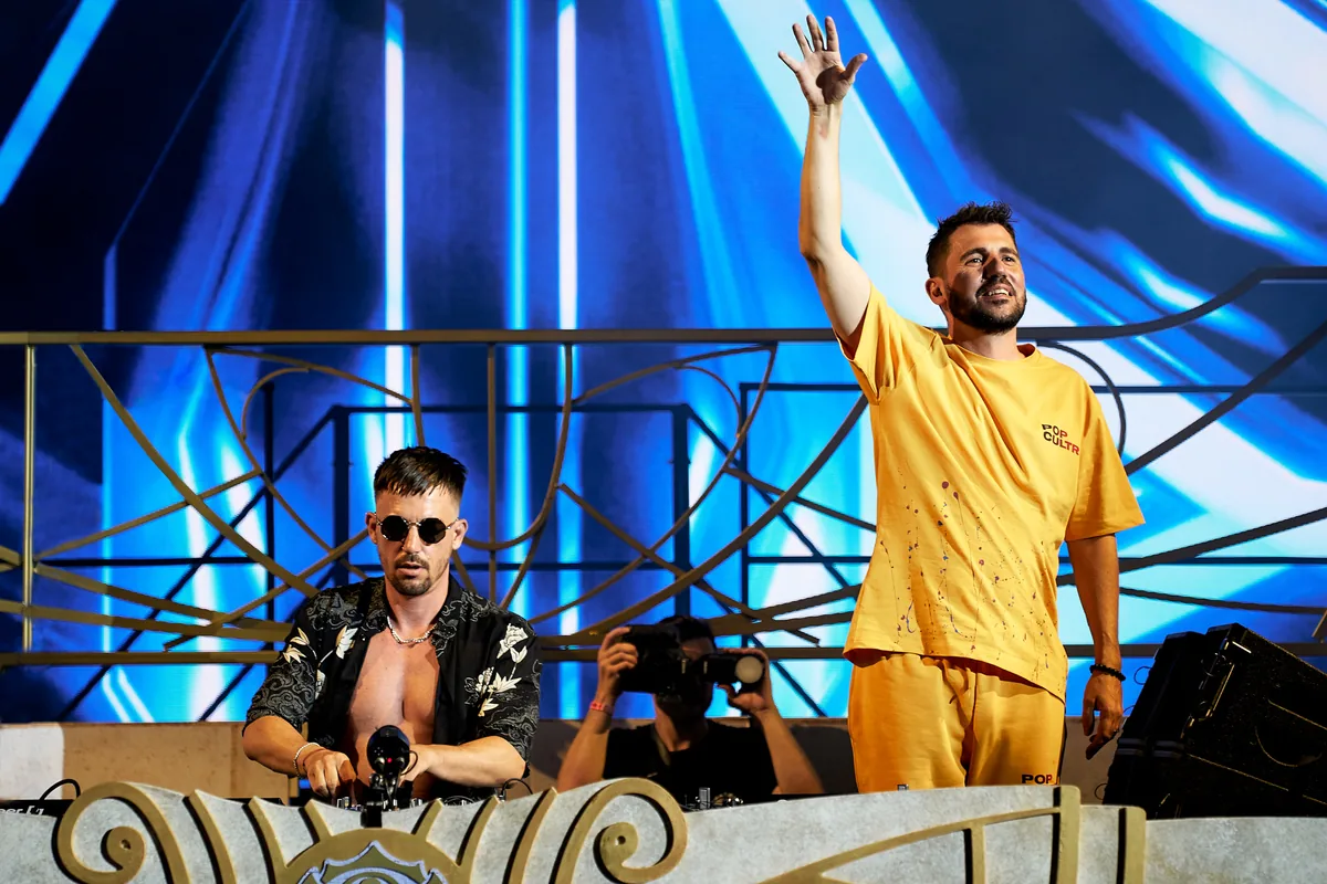 Dimitri Vegas and Like Mike behind the decks at Ushuaia Ibiza, one with a raised hand, in front of a blue video wall