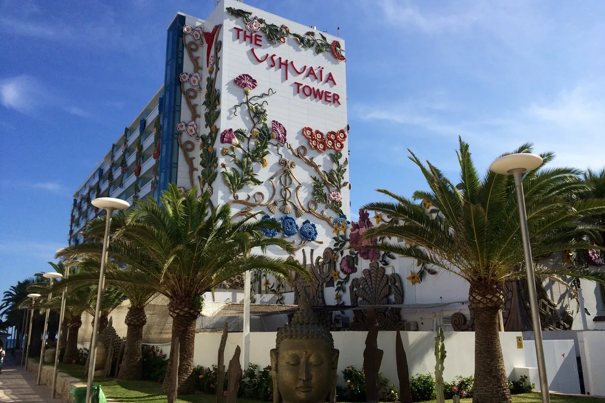 The white Ushuaia Tower of the beach hotel with flower sculptures on its wall, palm trees and a stone head in the foreground